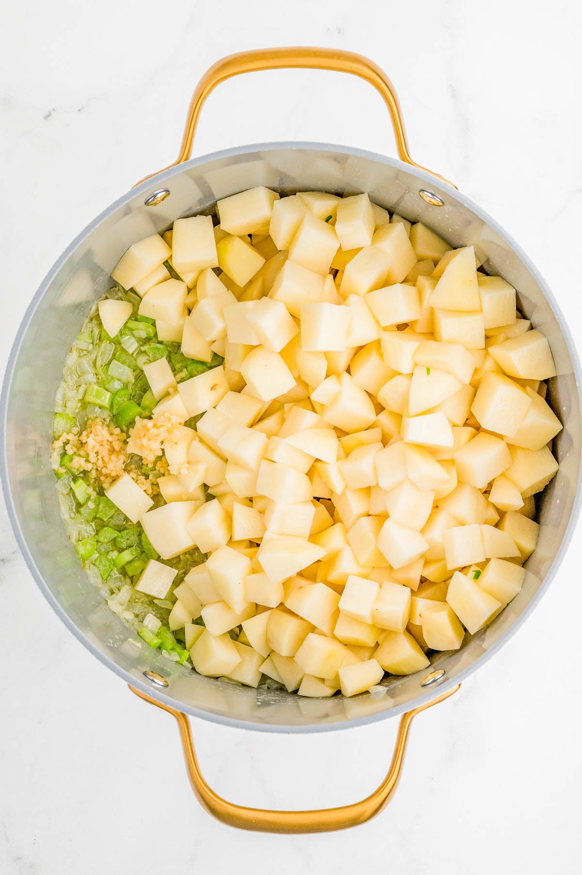 A pot contains chopped potatoes, celery, onion, and minced garlic, ready to be cooked.