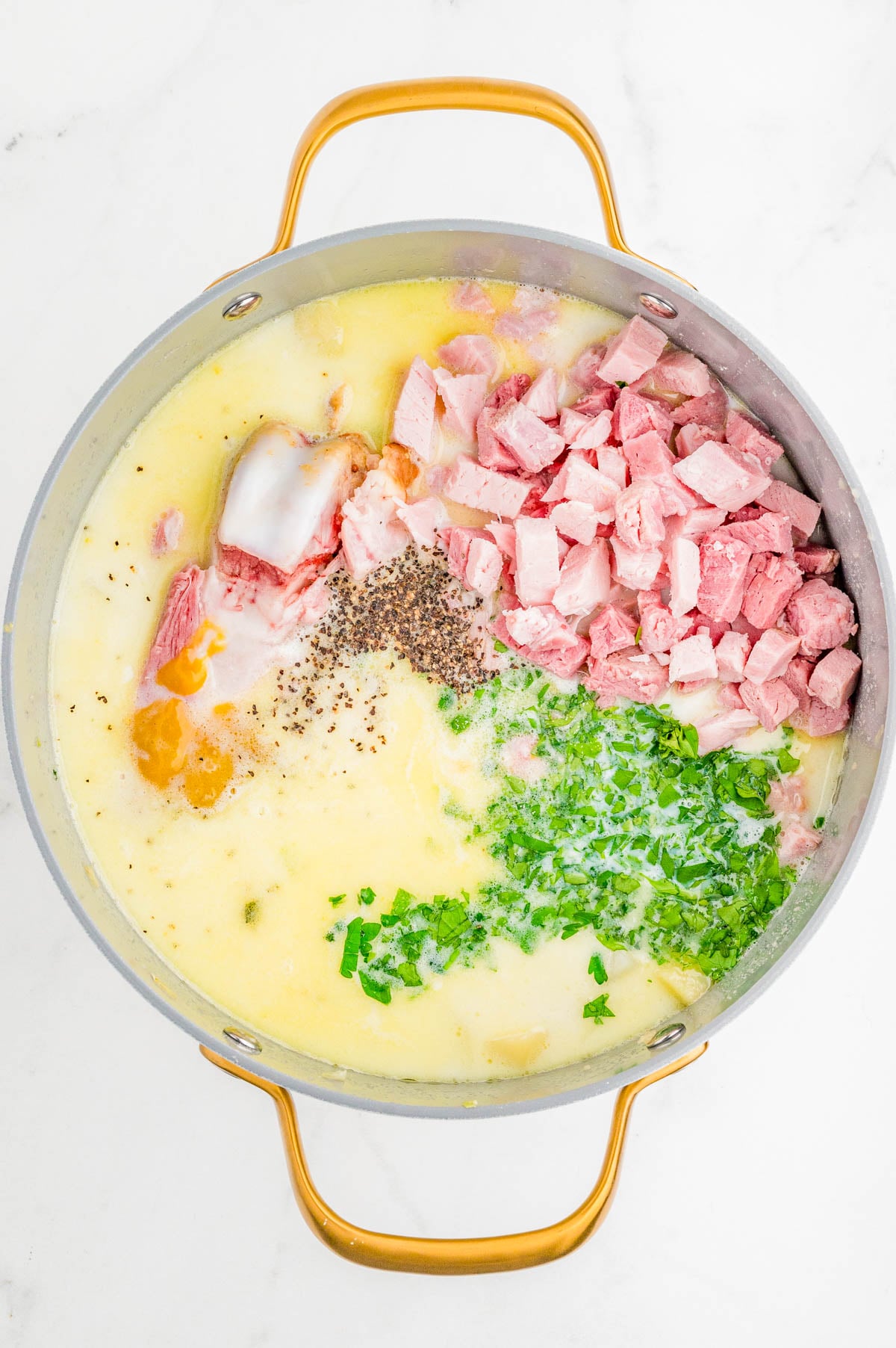 A pot filled with chopped ham, potatoes, milk, herbs, black pepper, and other soup ingredients, ready to be mixed.
