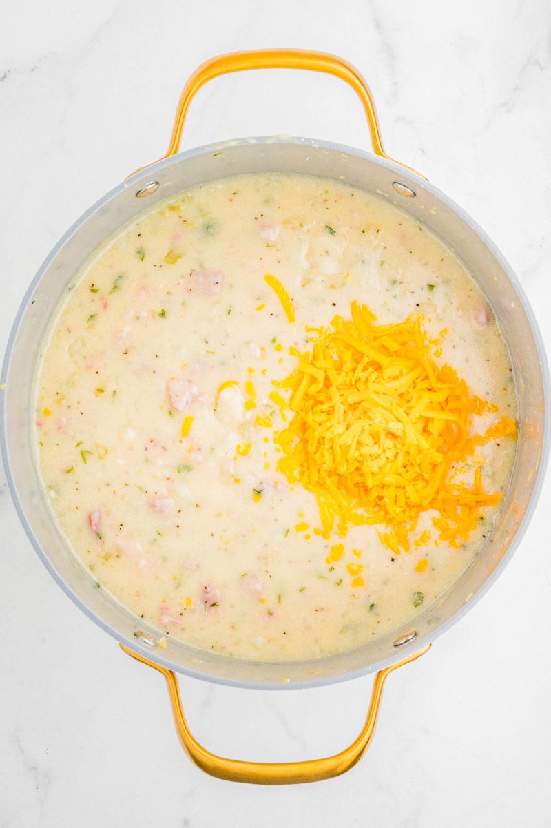 A pot of creamy soup with chopped herbs and bacon, topped with a pile of shredded cheddar cheese, sits on a white marble surface.