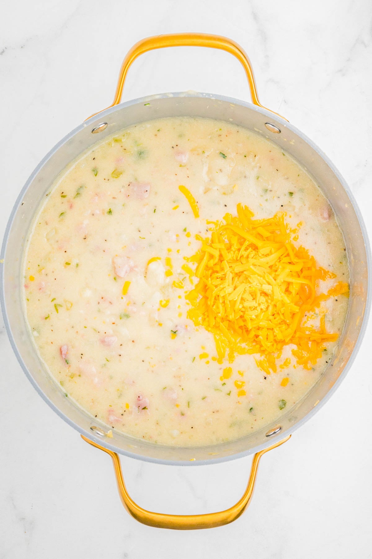 A pot of creamy soup with chopped herbs and bacon, topped with a pile of shredded cheddar cheese, sits on a white marble surface.