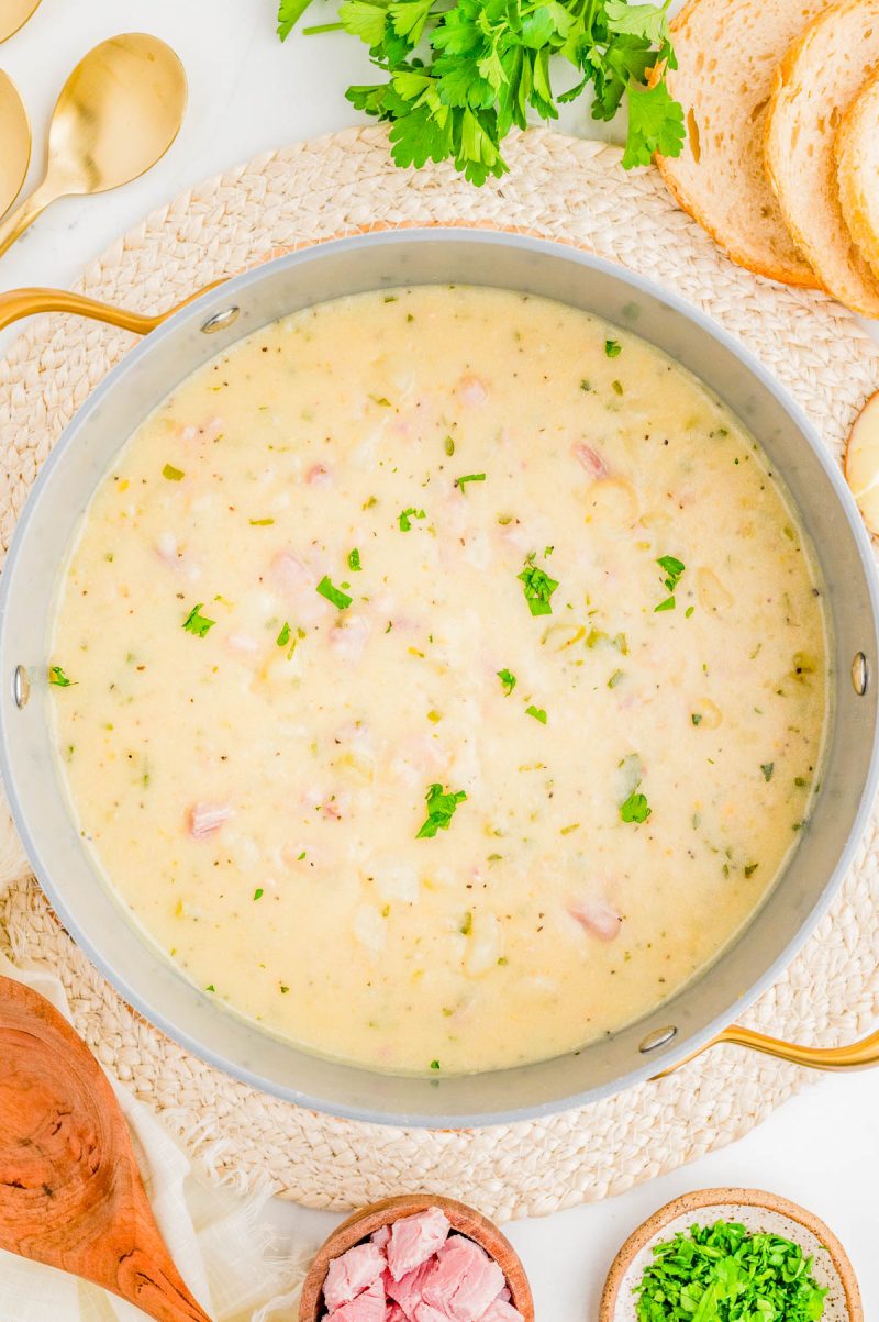 A pot of creamy soup garnished with chopped parsley, surrounded by slices of bread, fresh herbs, and bowls of diced ham and parsley on a light woven placemat.
