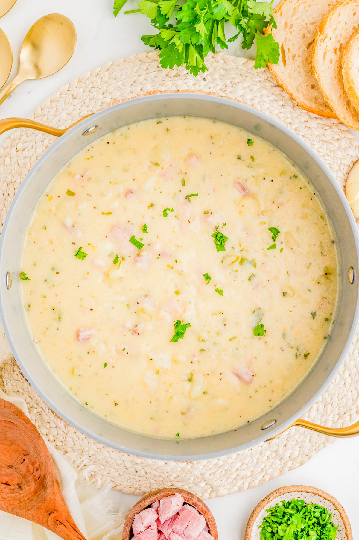 A pot of creamy soup garnished with chopped parsley, surrounded by slices of bread, fresh herbs, and bowls of diced ham and parsley on a light woven placemat.