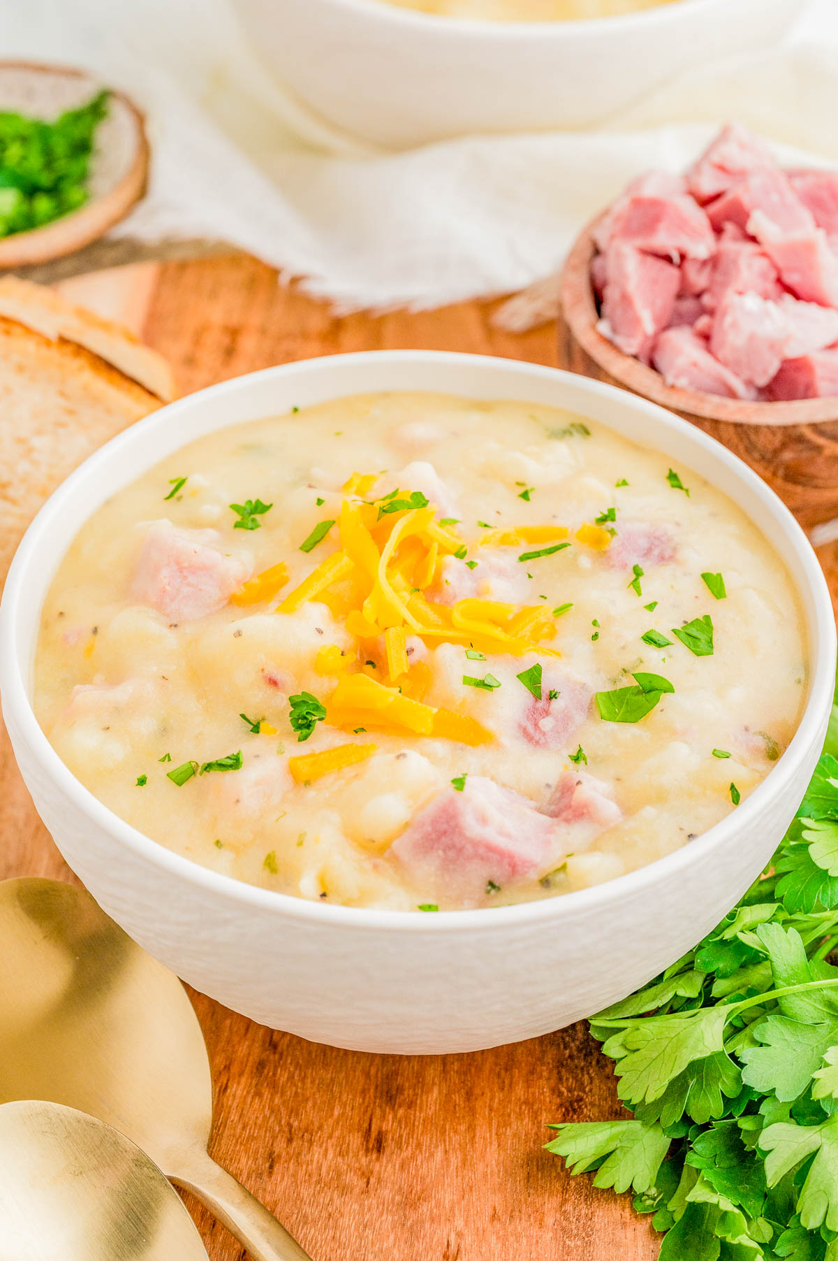 A bowl of creamy potato soup with ham, garnished with shredded cheese and chopped parsley, sits on a wooden surface next to a spoon, fresh parsley, and a bowl of diced ham.