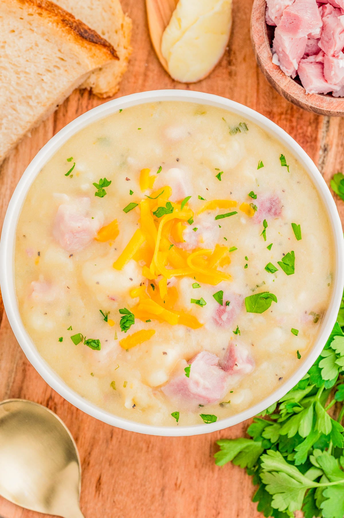 A bowl of creamy soup with ham, potatoes, shredded cheese, and chopped parsley, surrounded by sliced bread, parsley, and a bowl of diced ham on a wooden surface.