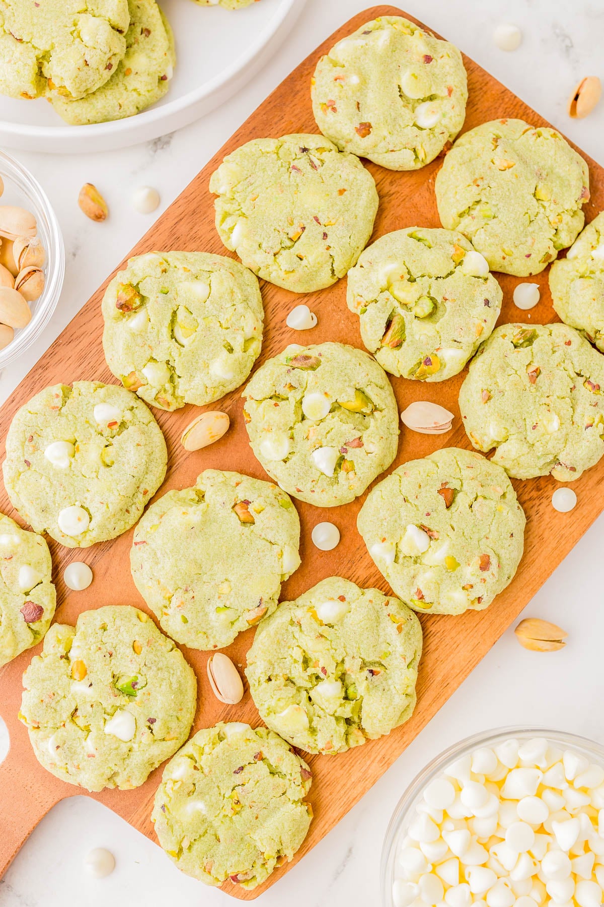 A wooden board with several green cookies containing white chocolate chips and pistachio pieces, surrounded by bowls of pistachios and white chocolate chips.