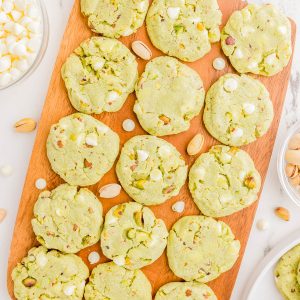 A wooden board with 16 green pistachio cookies topped with white chocolate chips, surrounded by scattered pistachios and a bowl of white chocolate chips.