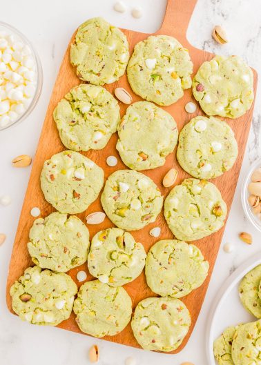 A wooden board with 16 green pistachio cookies topped with white chocolate chips, surrounded by scattered pistachios and a bowl of white chocolate chips.
