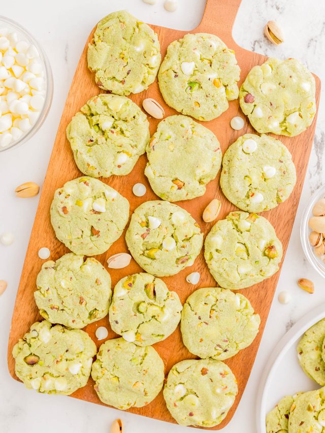 A wooden board with 16 green pistachio cookies topped with white chocolate chips, surrounded by scattered pistachios and a bowl of white chocolate chips.