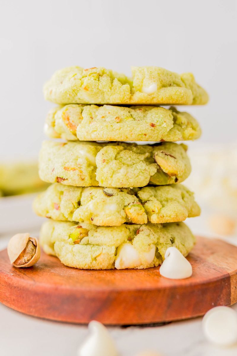 A stack of green pistachio cookies with white chocolate chips on a wooden board, surrounded by pistachios and white chocolate chips.