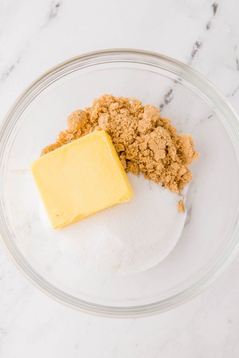 A glass bowl containing a stick of butter, brown sugar, and granulated white sugar on a white marble surface.