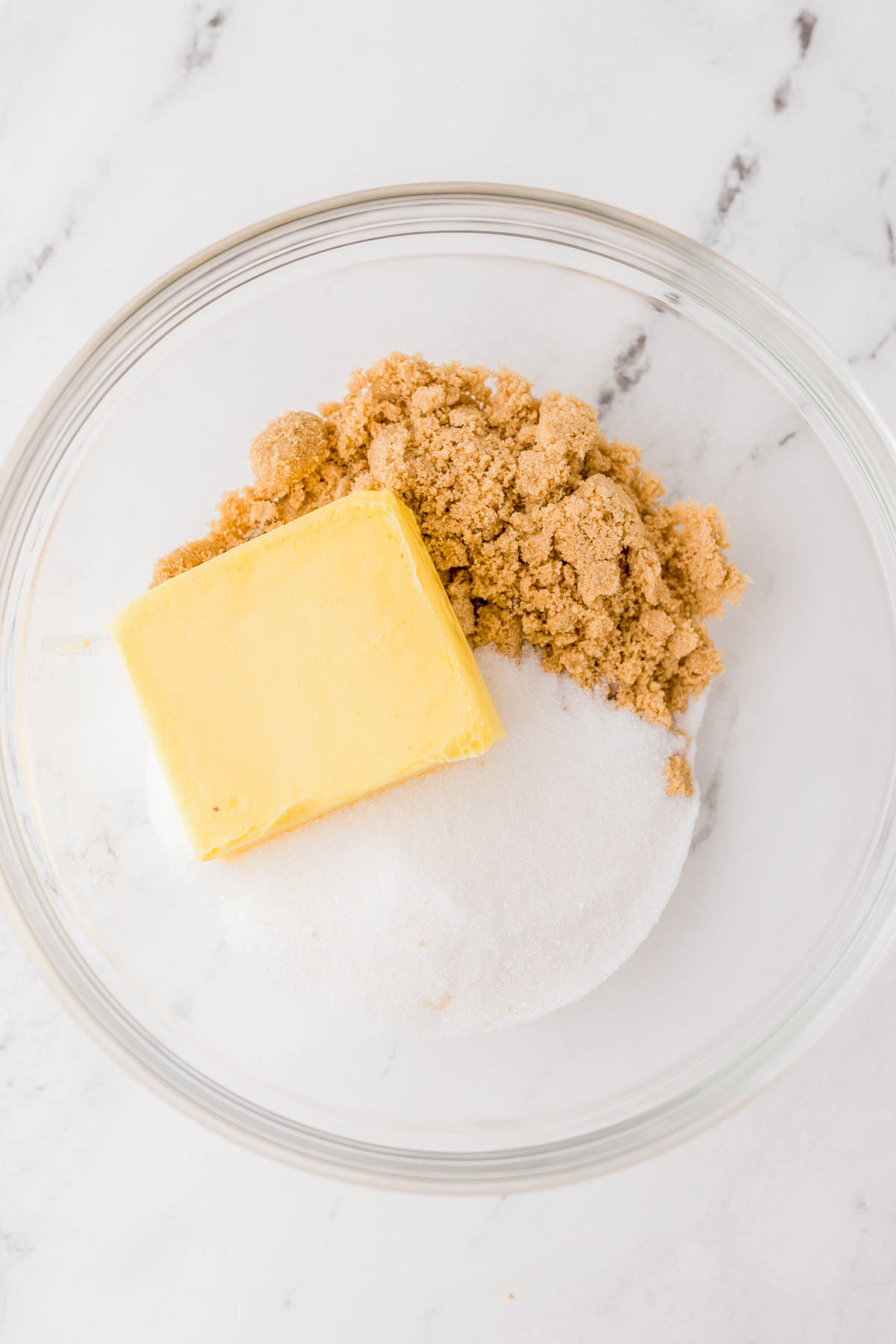 A glass bowl containing a stick of butter, brown sugar, and granulated white sugar on a white marble surface.
