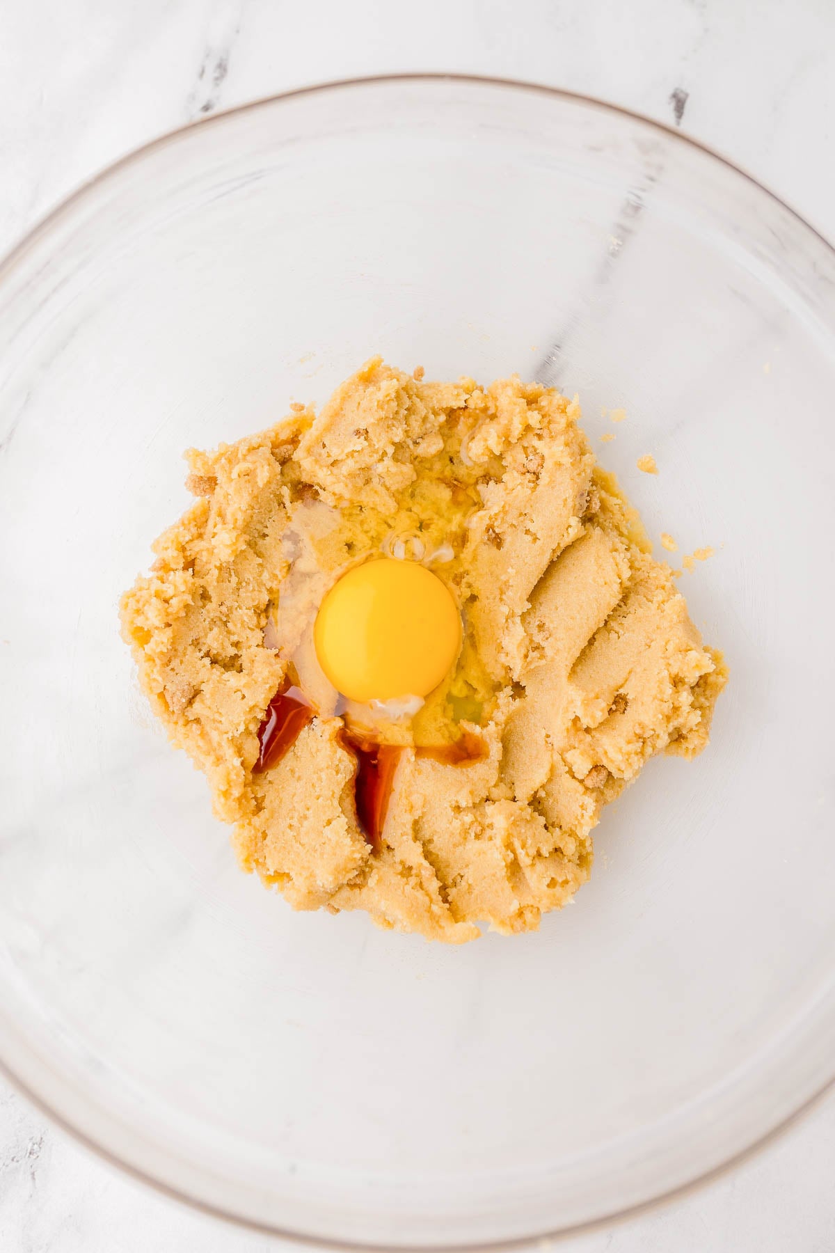 A glass bowl containing creamed butter and sugar with an egg and vanilla extract added in the center, ready to be mixed.