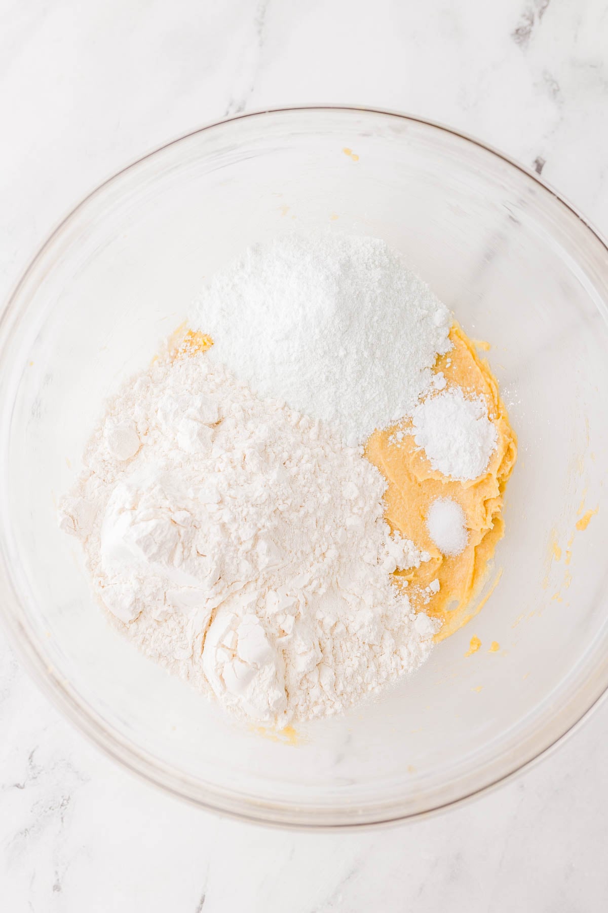 A glass mixing bowl containing flour, sugar, baking powder, and a yellow batter mixture on a white marble surface.