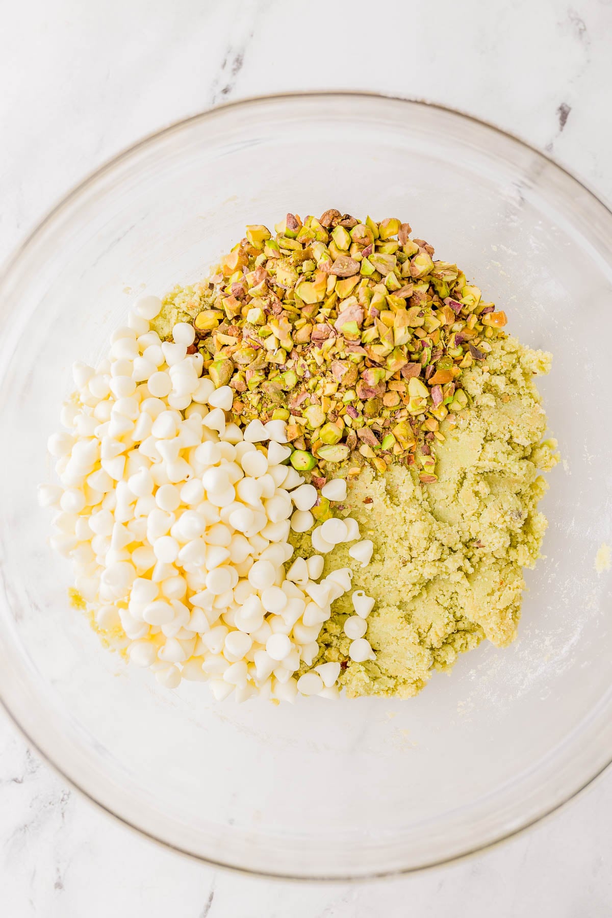 A glass bowl containing white chocolate chips, chopped pistachios, and cookie dough mixture on a marble surface.
