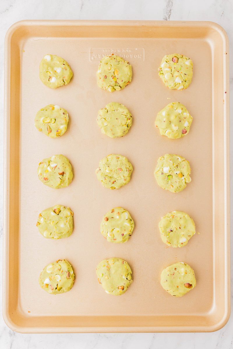 A baking sheet with fifteen unbaked cookie dough pieces containing white chips and chopped nuts, evenly spaced in rows.