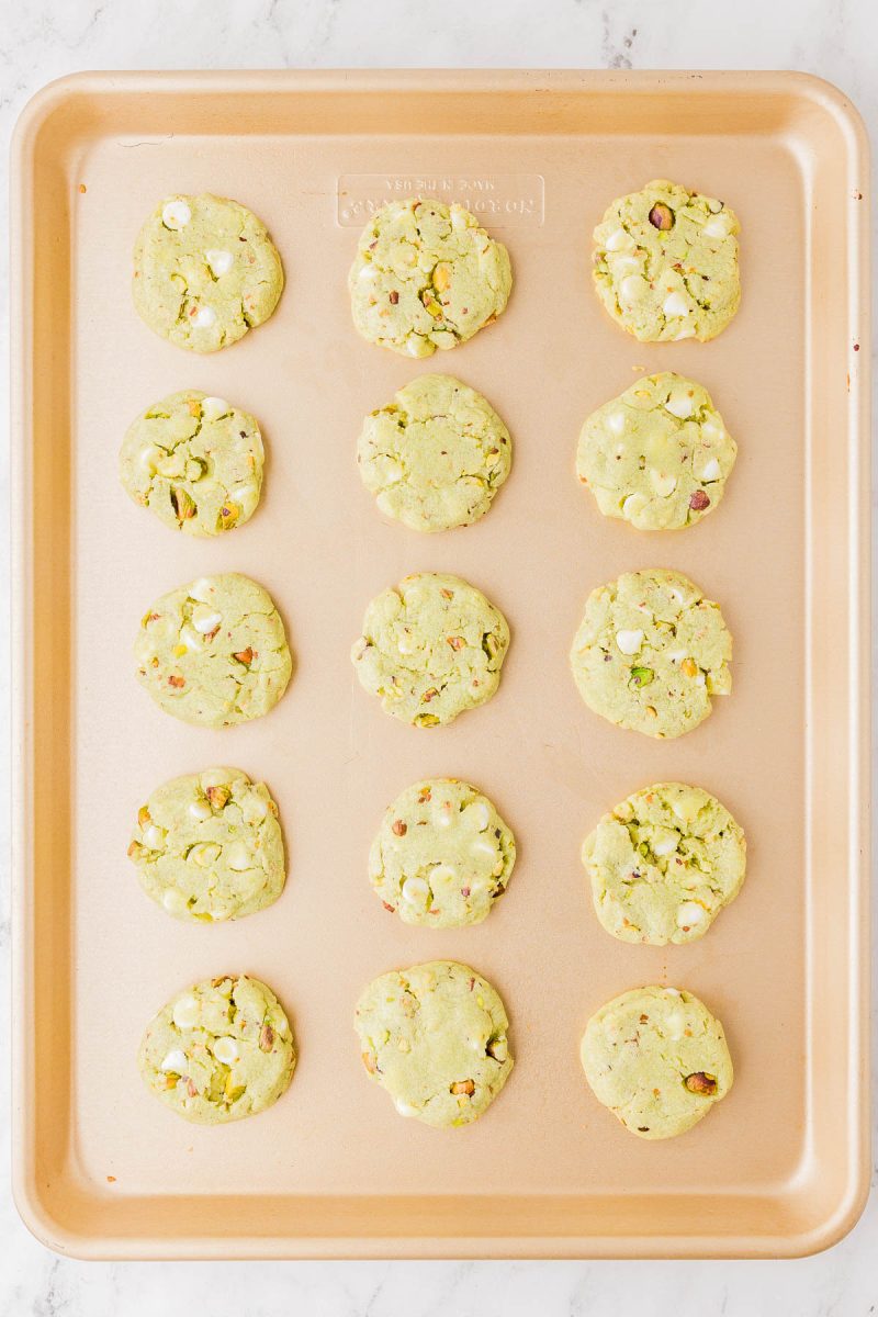 A baking sheet with fifteen round cookies containing white chocolate chips and chopped pistachios, arranged in rows on a light surface.