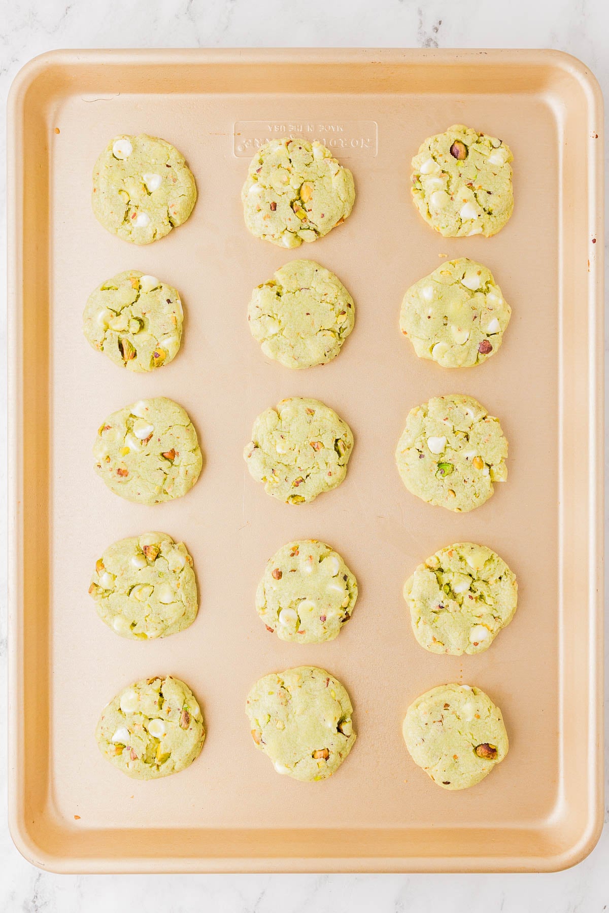 A baking sheet with fifteen round cookies containing white chocolate chips and chopped pistachios, arranged in rows on a light surface.