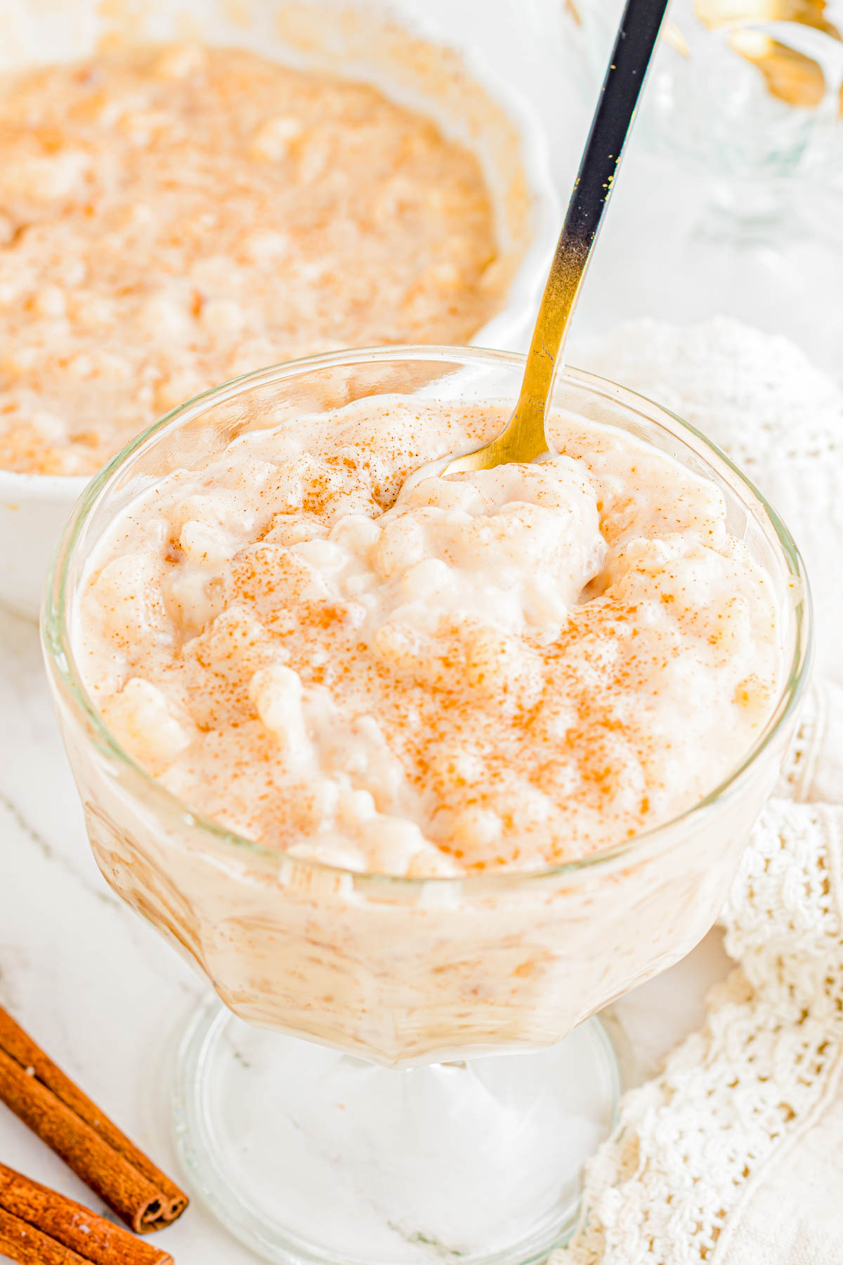 A glass dessert dish filled with creamy rice pudding, topped with a sprinkle of cinnamon, with a spoon in the pudding. A bowl of extra pudding and cinnamon sticks are in the background.