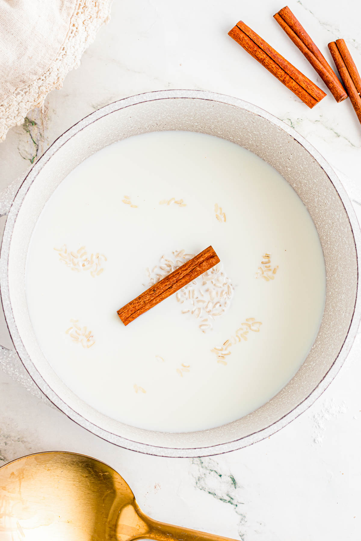 A white pot filled with milk, uncooked rice, and a cinnamon stick on a marble countertop, with a gold spoon and extra cinnamon sticks nearby.