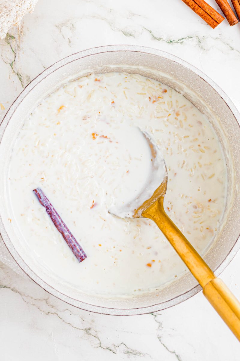 A pot of rice pudding with a cinnamon stick, partially stirred with a gold ladle, sits on a white marble surface.