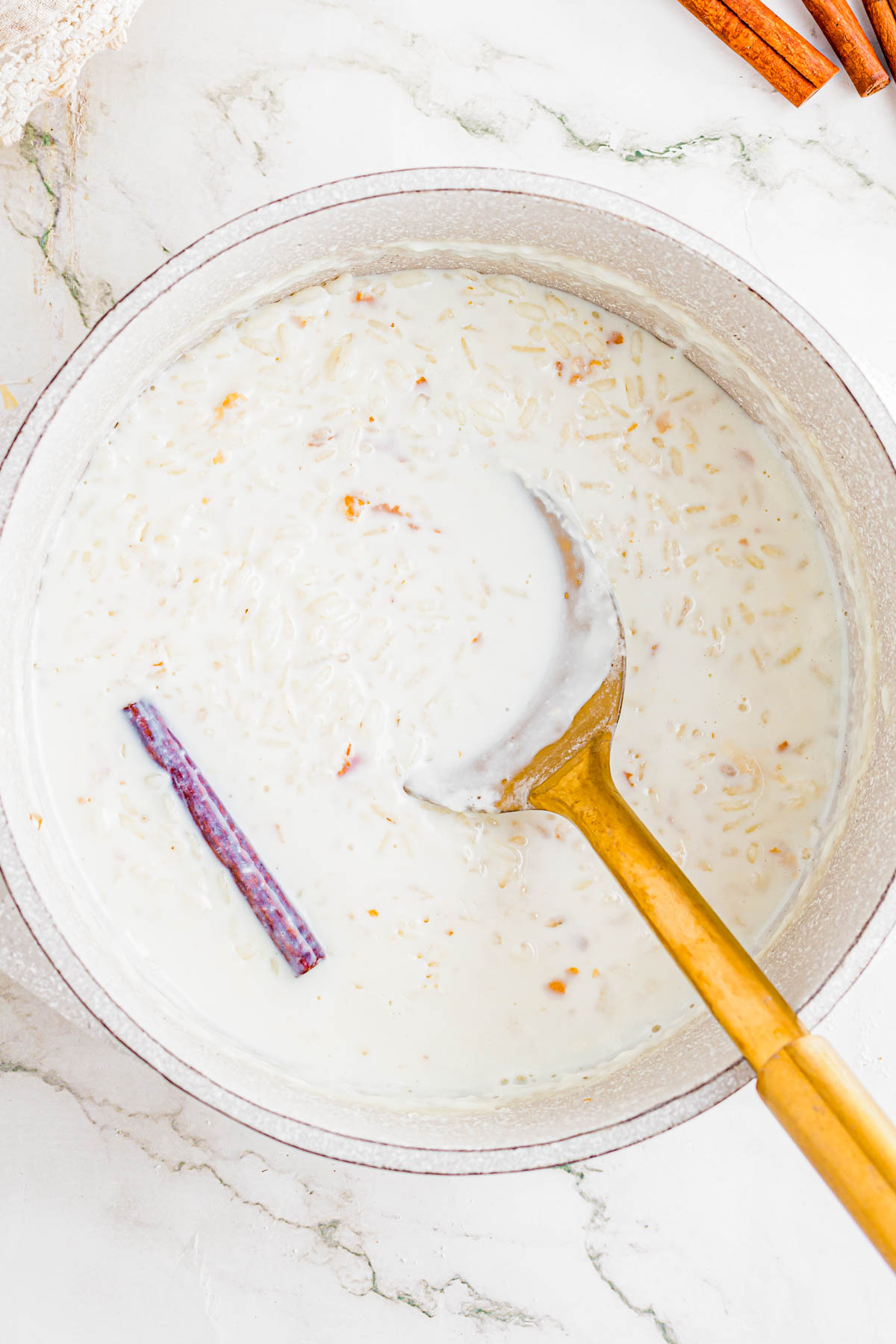 A pot of rice pudding with a cinnamon stick, partially stirred with a gold ladle, sits on a white marble surface.