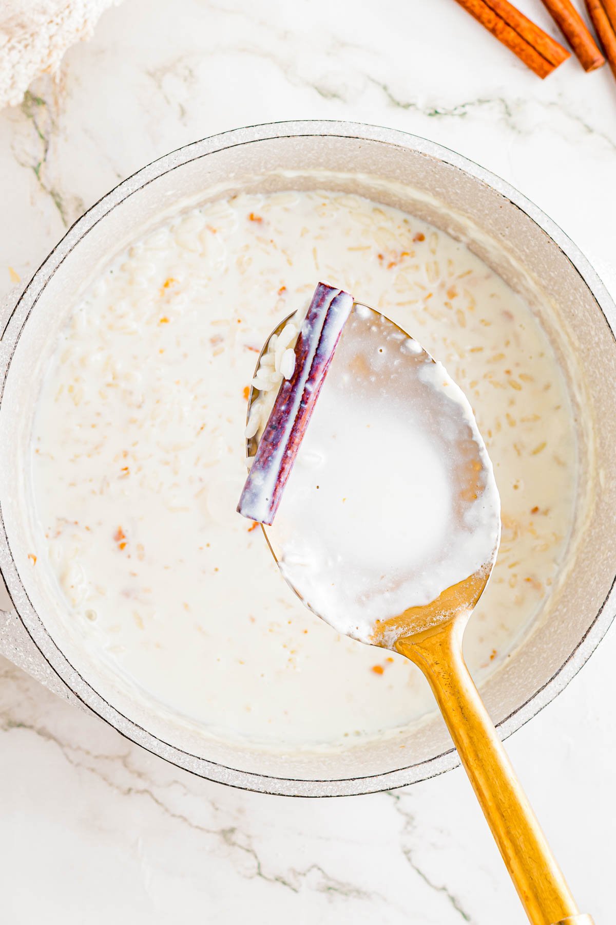 A pot of rice pudding in milk with a gold spoon holding a cinnamon stick and grains of rice above the mixture.