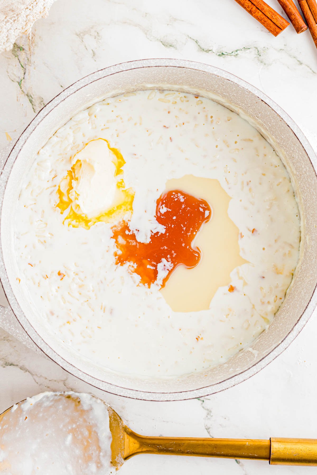 A pot of rice pudding mixture with vanilla extract, egg yolk, and cream added but not yet mixed, next to a golden spoon on a marble countertop.