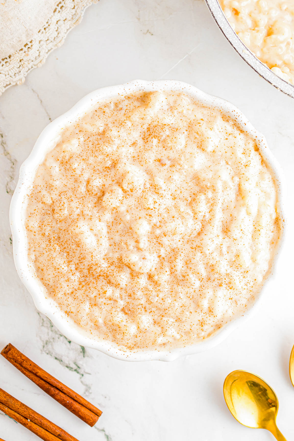 A white bowl filled with rice pudding, sprinkled with ground cinnamon, sits on a white surface near golden spoons and cinnamon sticks.