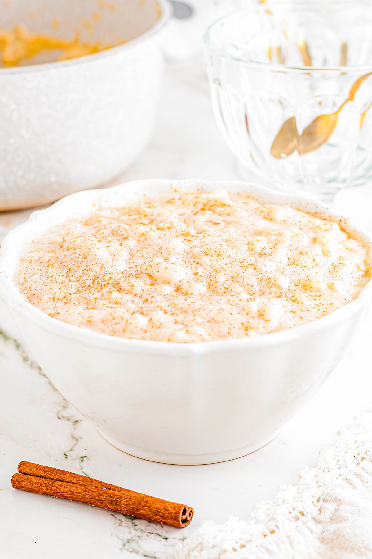 A white bowl filled with rice pudding, topped with a sprinkle of cinnamon, sits on a marble surface near a cinnamon stick.
