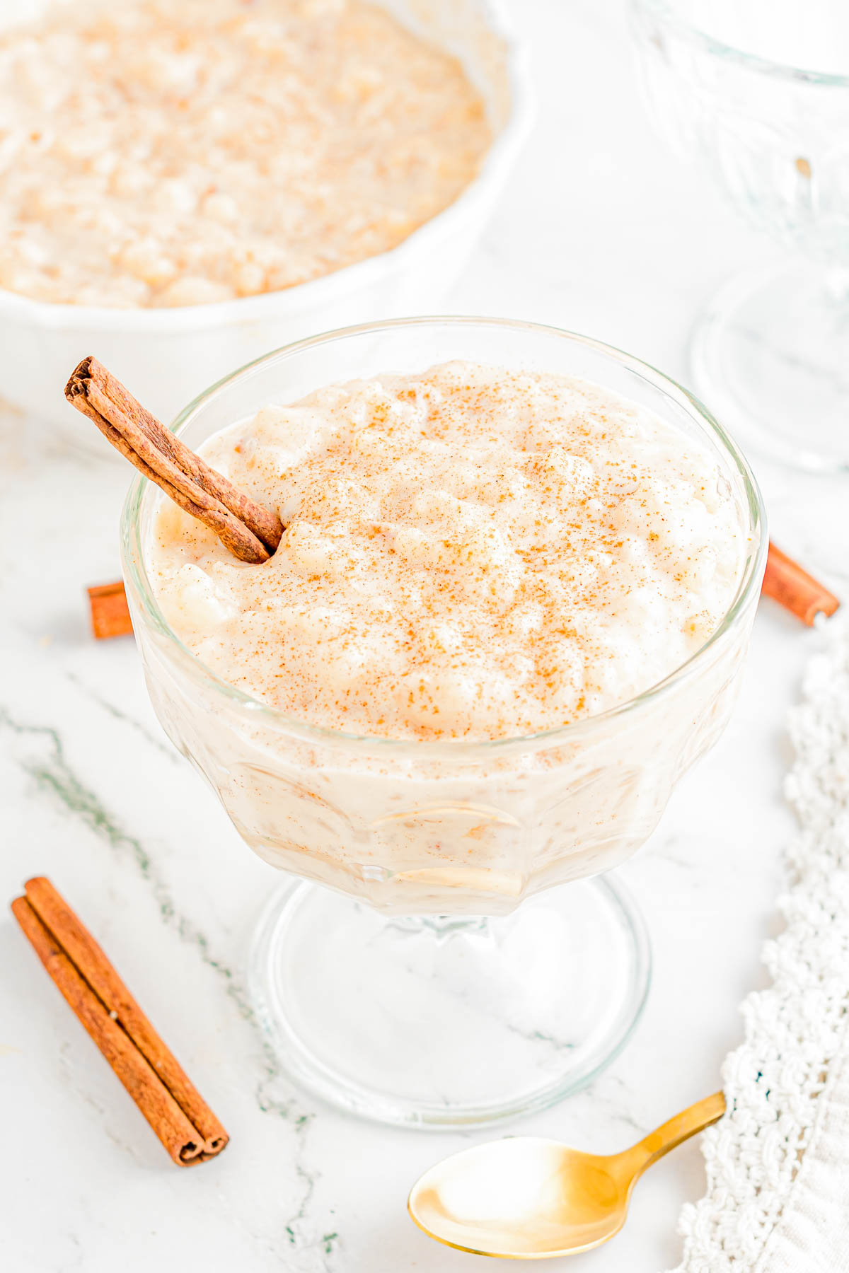 A glass dessert dish filled with rice pudding, topped with ground cinnamon and a cinnamon stick, sits on a white surface with cinnamon sticks and a gold spoon nearby.