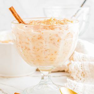 A glass dish filled with creamy rice pudding, topped with ground cinnamon and a cinnamon stick, with a gold spoon and extra cinnamon sticks beside it.