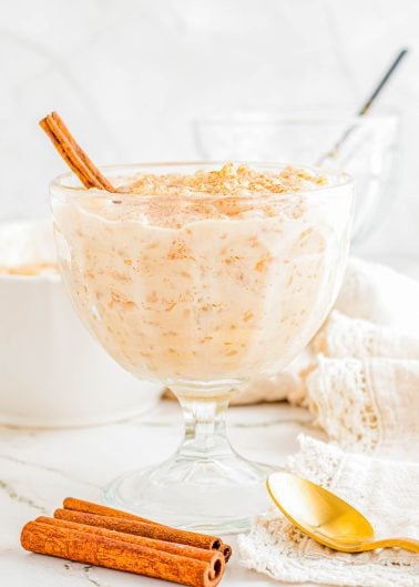 A glass dish filled with creamy rice pudding, topped with ground cinnamon and a cinnamon stick, with a gold spoon and extra cinnamon sticks beside it.