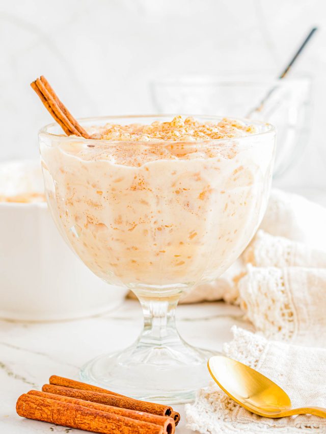 A glass dish filled with creamy rice pudding, topped with ground cinnamon and a cinnamon stick, with a gold spoon and extra cinnamon sticks beside it.