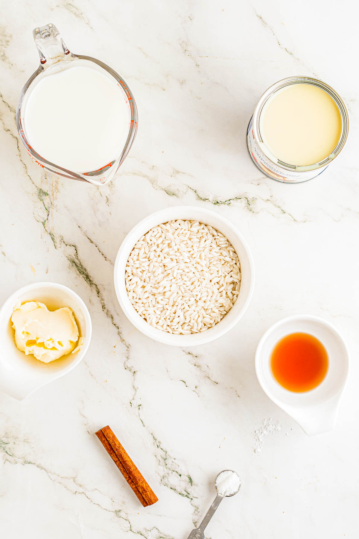Ingredients for rice pudding laid out on a white marble surface: a bowl of rice, milk, sweetened condensed milk, butter, vanilla extract, cinnamon stick, and measuring spoon with salt.