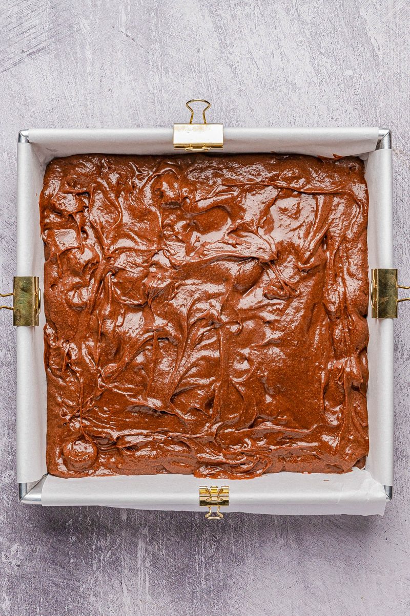 A rectangular baking pan lined with parchment paper holds unbaked chocolate brownie batter, secured with metal clips on the edges.