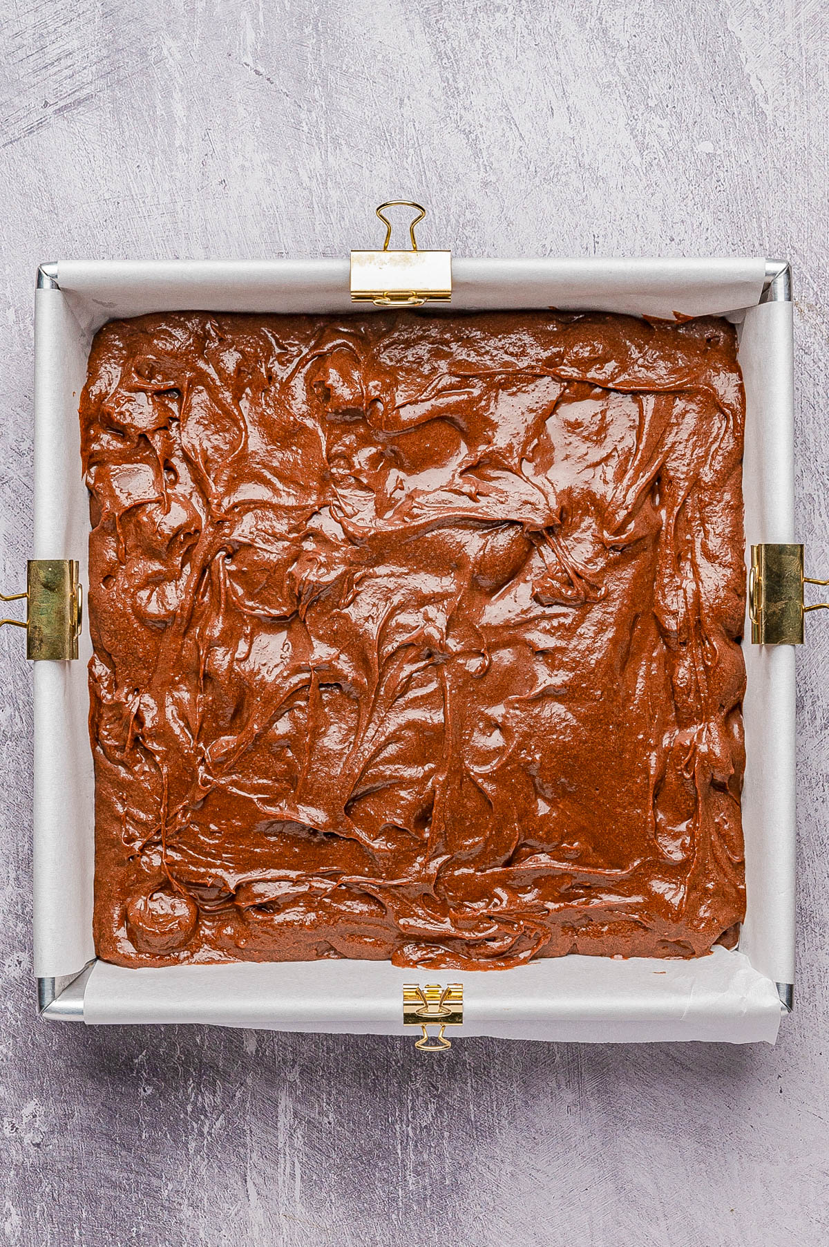 A rectangular baking pan lined with parchment paper holds unbaked chocolate brownie batter, secured with metal clips on the edges.