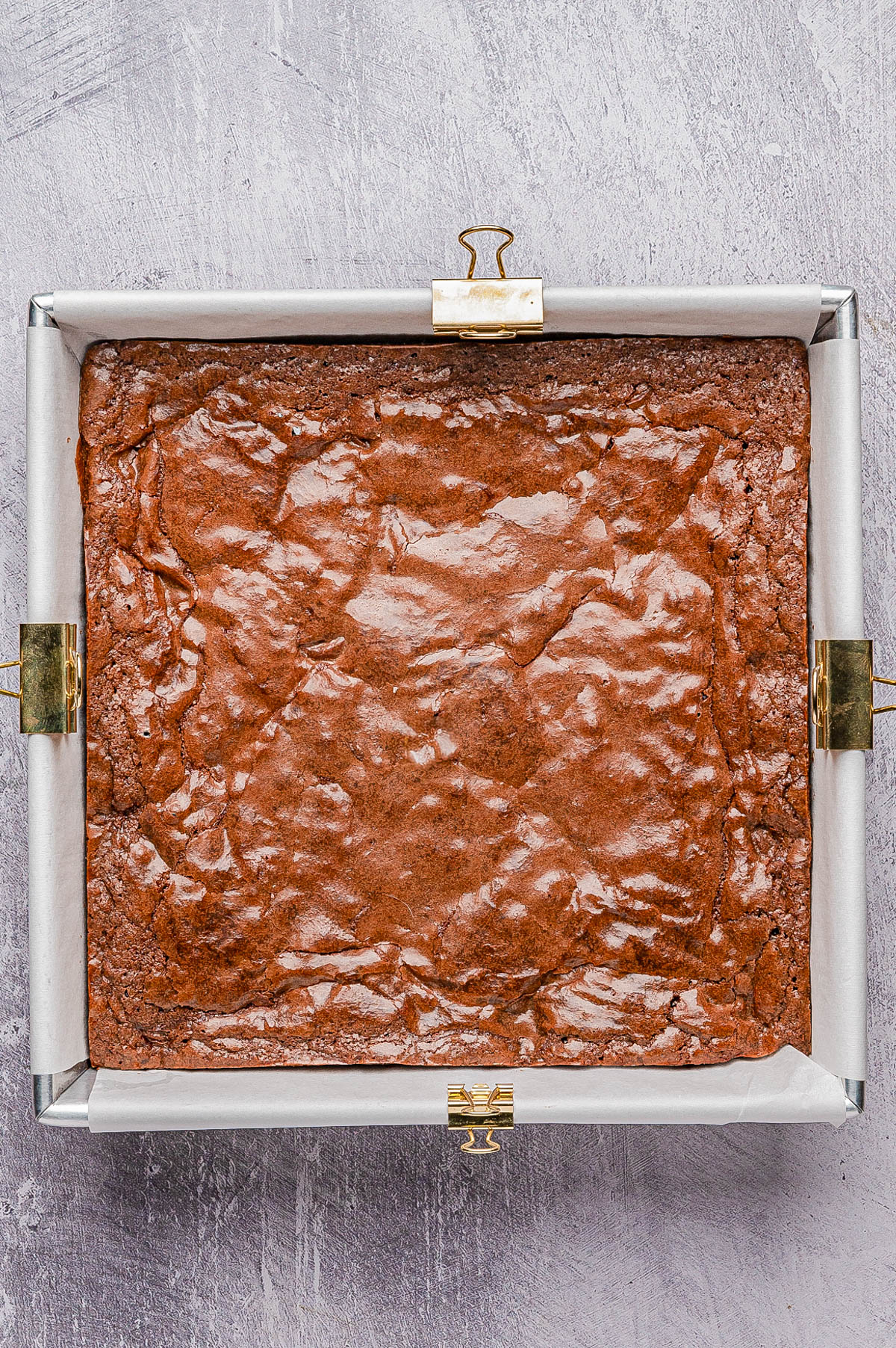 A square pan lined with parchment paper holds a freshly baked batch of brownies with a shiny, crinkled top.