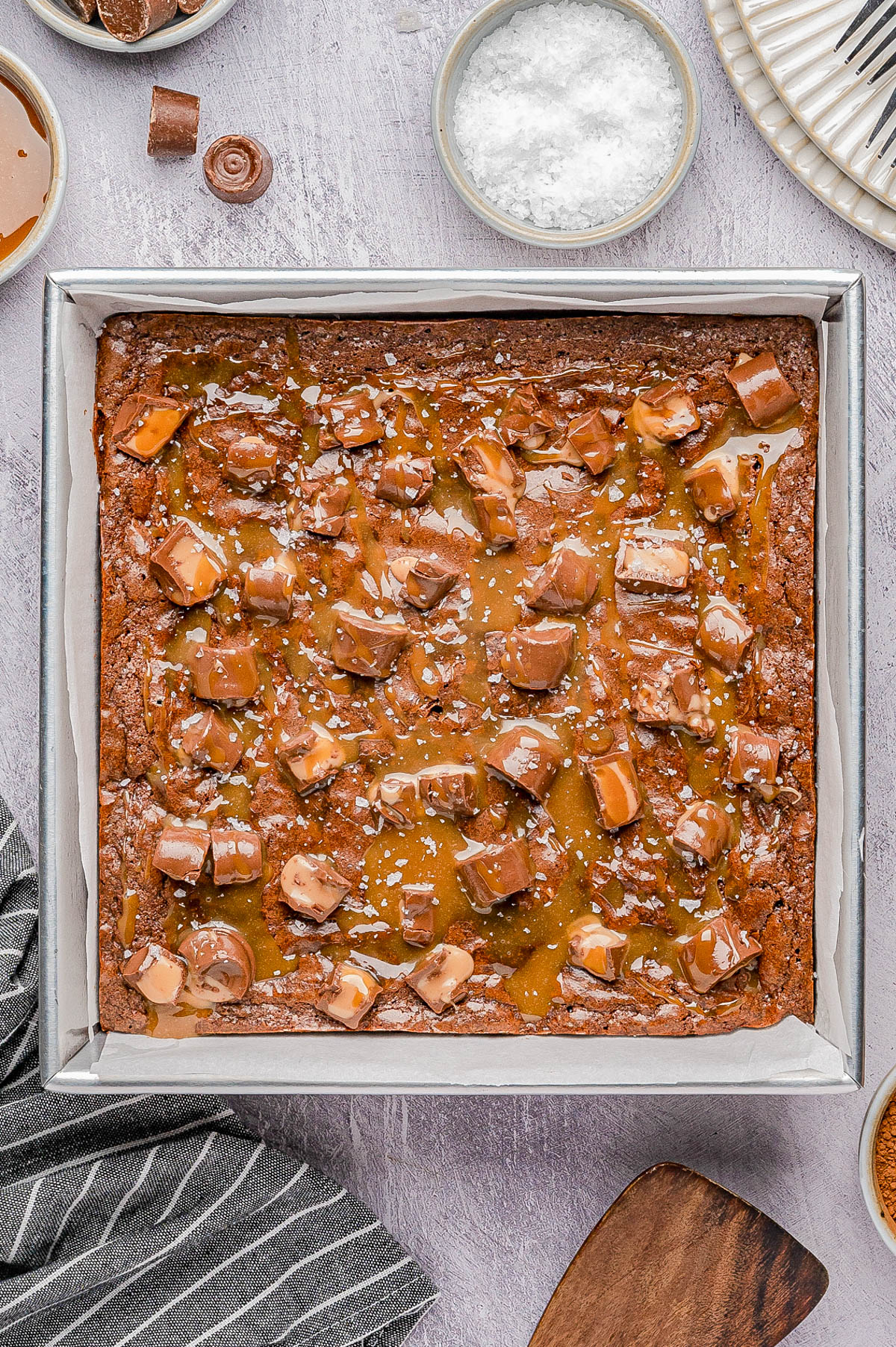 A tray of brownies topped with caramel drizzle, chopped chocolate pieces, and sprinkled sea salt, sitting on a parchment-lined pan.