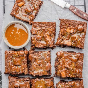 Nine chocolate brownies topped with caramel sauce are arranged on parchment paper over a cooling rack, with a bowl of caramel and a knife nearby.