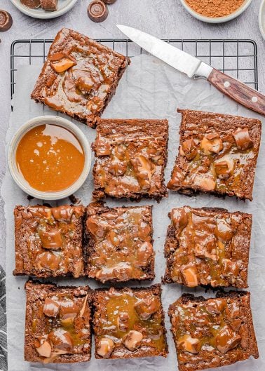 Nine chocolate brownies topped with caramel sauce are arranged on parchment paper over a cooling rack, with a bowl of caramel and a knife nearby.