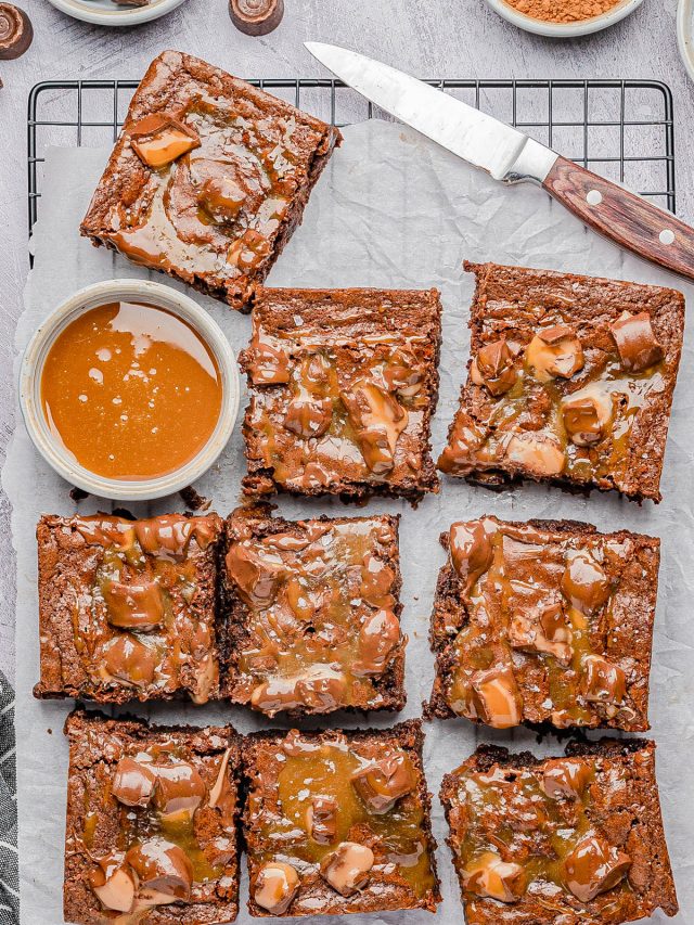 Nine chocolate brownies topped with caramel sauce are arranged on parchment paper over a cooling rack, with a bowl of caramel and a knife nearby.