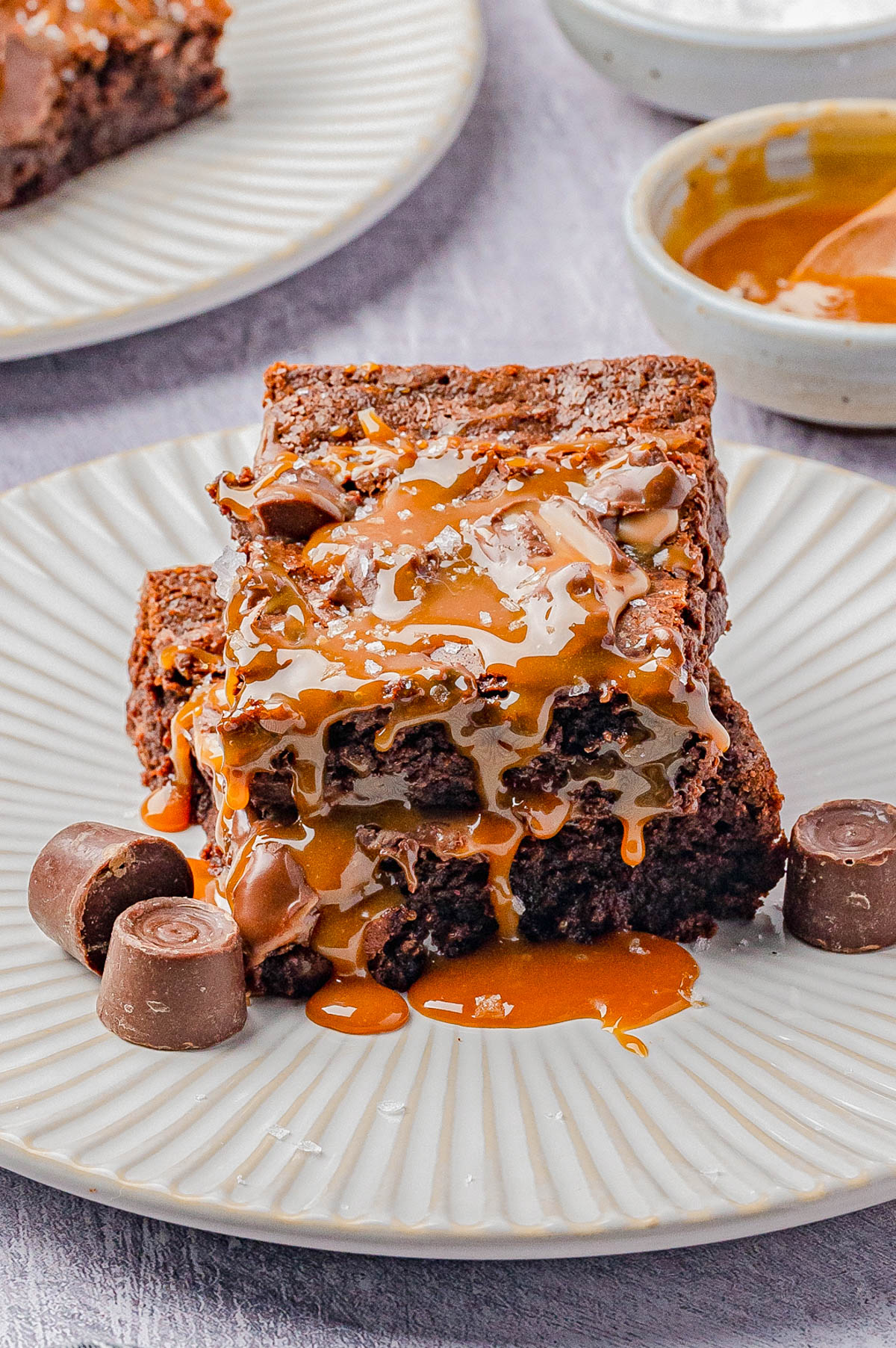 Two chocolate brownies stacked on a plate, drizzled with caramel sauce, with pieces of caramel chocolate candy beside them.
