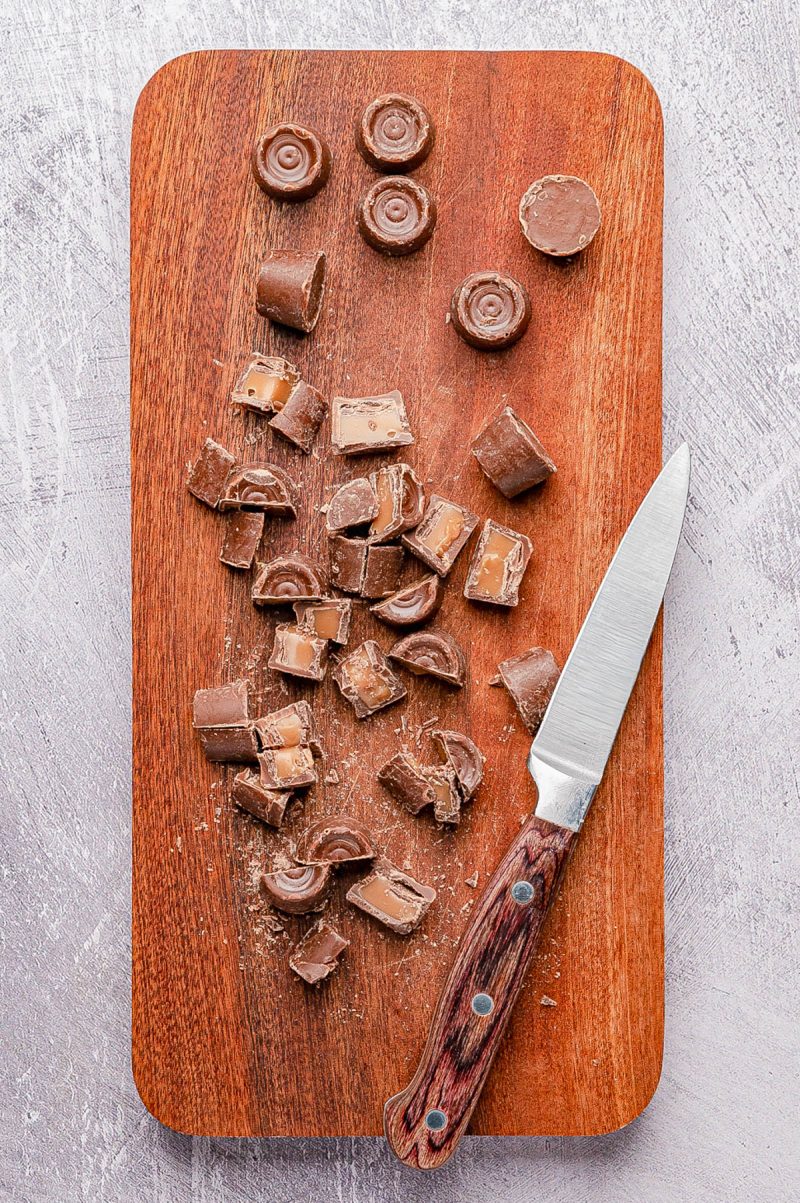 A wooden cutting board with a knife and several pieces of sliced chocolate candy, some with caramel filling visible.