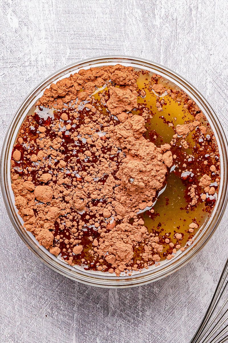 A glass bowl containing cocoa powder, honey, and oil on a textured light surface, ready to be mixed.