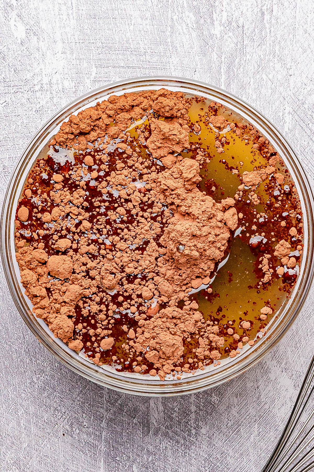 A glass bowl containing cocoa powder, honey, and oil on a textured light surface, ready to be mixed.