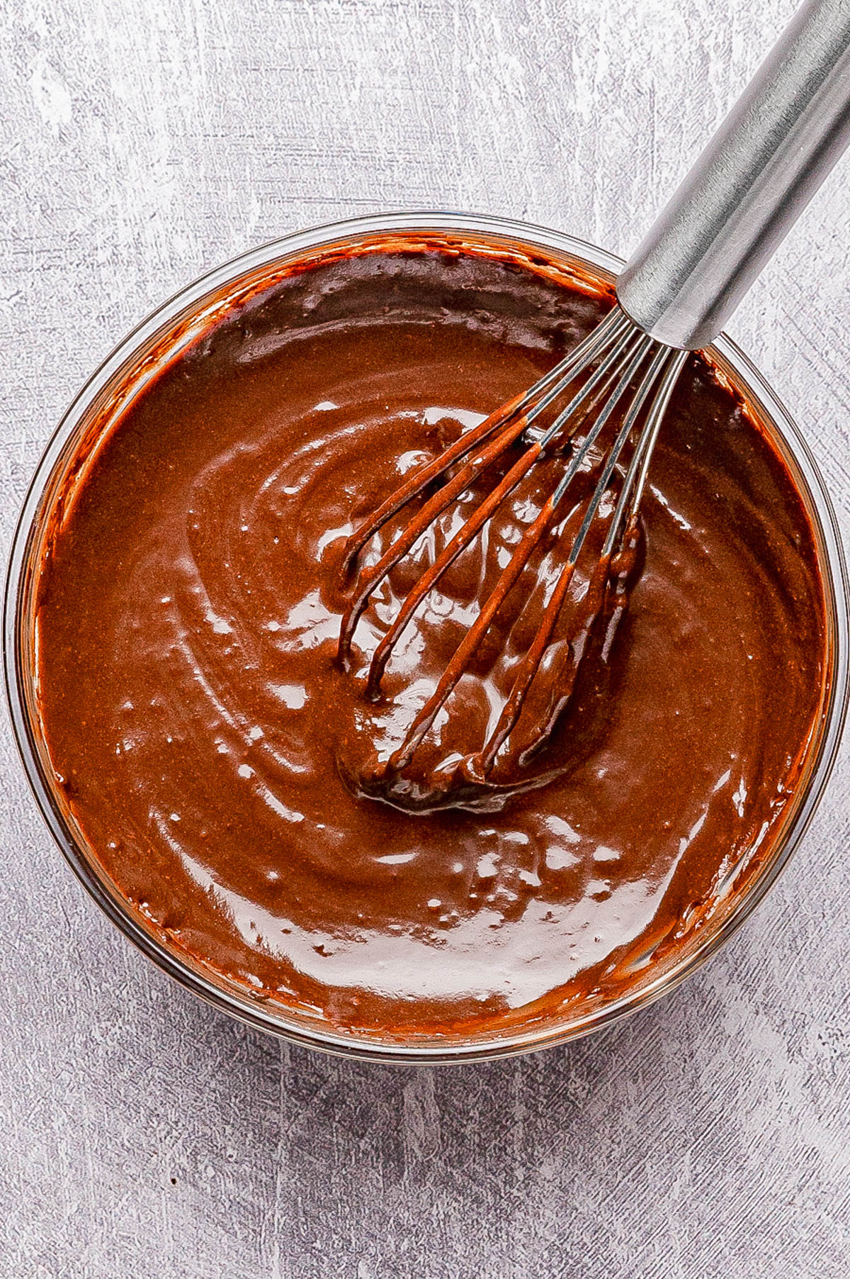 A metal whisk rests in a glass bowl filled with smooth chocolate cake batter on a textured light surface.