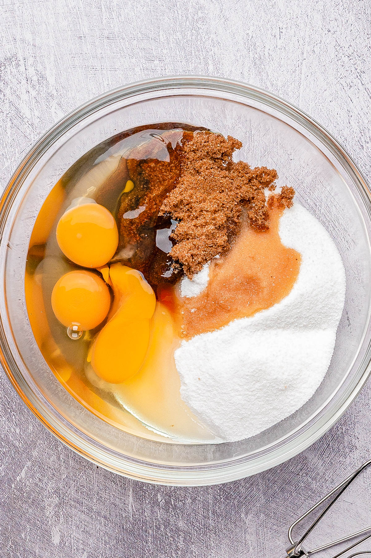 A glass bowl containing three raw eggs, brown sugar, white sugar, and vanilla extract, with a metal whisk nearby on a light-colored surface.