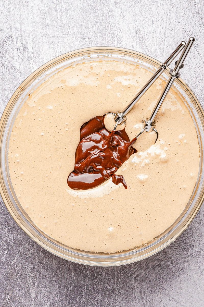 A glass bowl filled with light brown cake batter, topped with melted chocolate and two metal beaters resting inside.