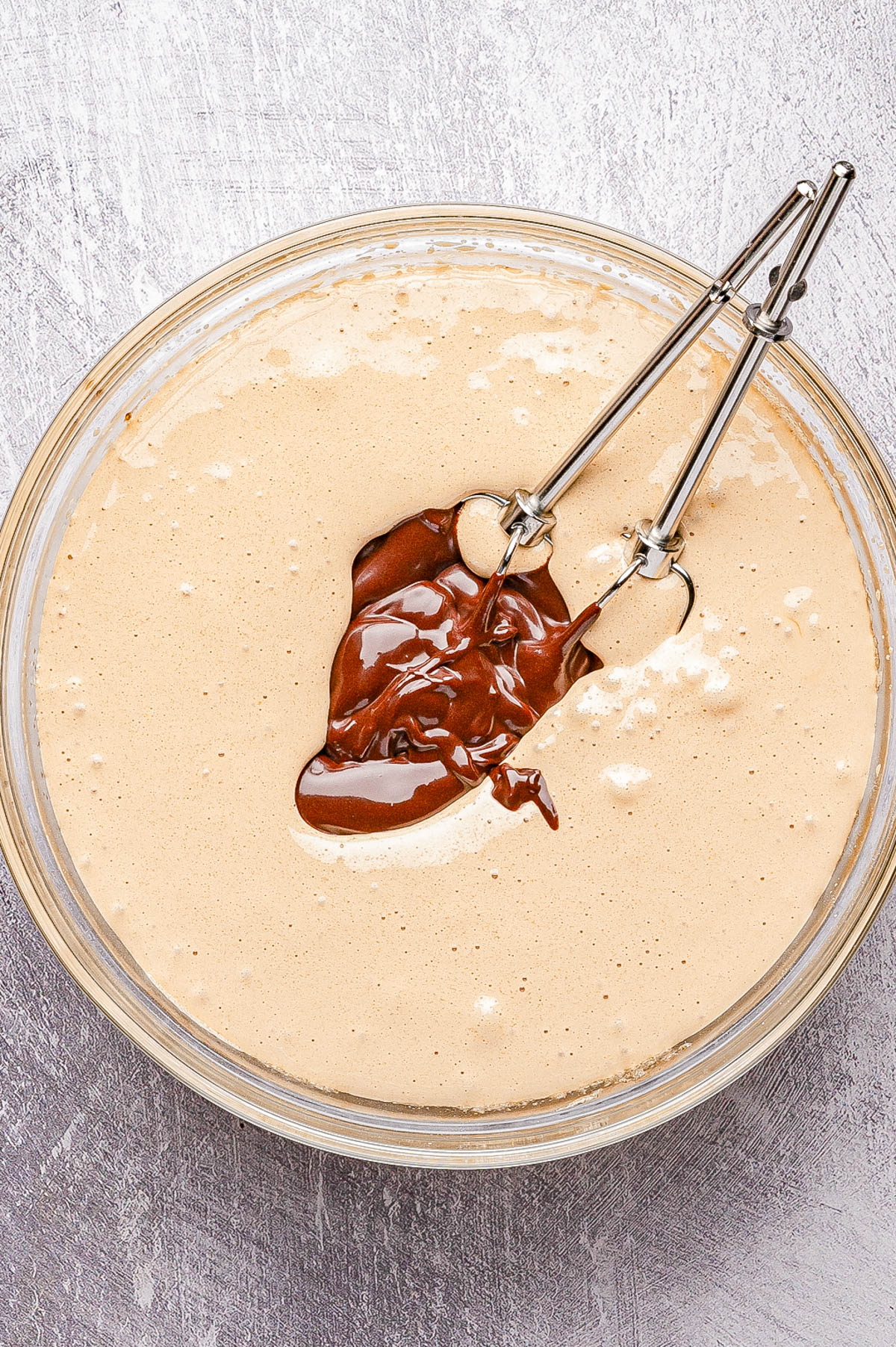 A glass bowl filled with light brown cake batter, topped with melted chocolate and two metal beaters resting inside.