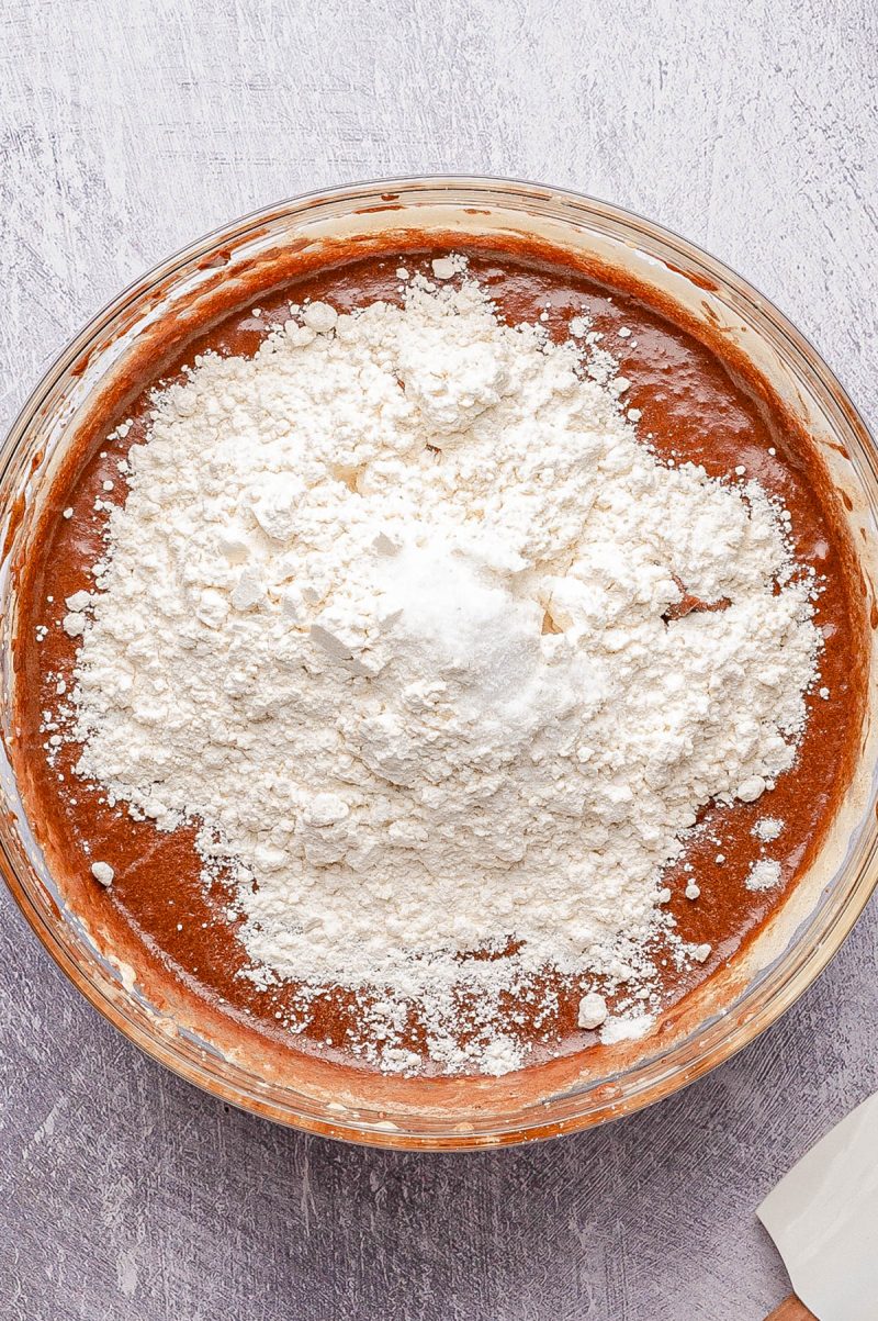 A glass bowl filled with chocolate batter topped with a mound of flour, ready to be mixed, sits on a light textured surface.